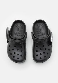 Sabots en mousse noire avec un design perforé, une sangle ajustable et une semelle intérieure texturée. La semelle présente une adhérence à motifs pour une meilleure traction.