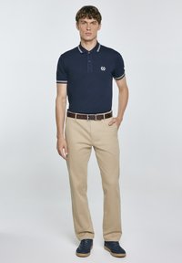 Tamsiai mėlynas polo marškinėlis su baltu apdailos kraštu, logotipas ant krūtinės; derinamas su kreminiais tiesiais keliais ir tamsiai mėlynais sportiniais batais. Lygus audinys, laisvas fasonas.