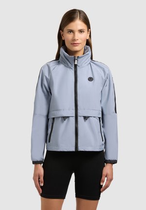 Žena v světle šedé bundě na zip s vysokým límcem a černých cyklistických šortkách na jednobarevném pozadí.