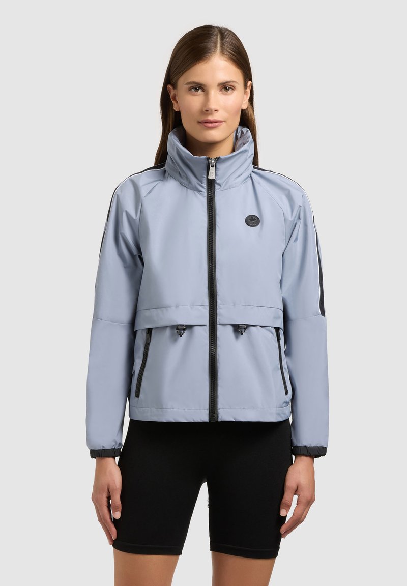 Femme portant une veste zippée gris clair à col montant et un cuissard de cyclisme noir, sur un fond uni.