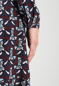Kleid mit mehrfarbigem Muster, das abstrakte Formen in Burgunderrot, Blau und Creme zeigt. Aus leichtem Stoff mit einer Knopfdetail am Ärmel.