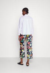 Weißes Langarmshirt mit Rückenpasse, kombiniert mit bunten, floral gemusterten Hosen auf marineblauem Grund und schwarzen Slippern.