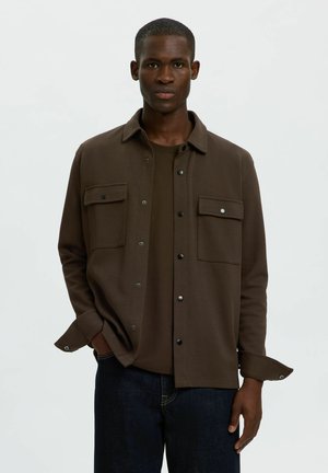 Junger Mann trägt ein dunkelbraunes, geknöpftes Overshirt mit Brusttaschen über einem passenden T-Shirt und dunkelblauen Jeans, steht mit einer Hand in der Tasche.