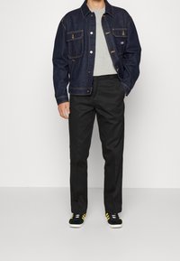 Homme portant une veste en jean bleu foncé, un t-shirt gris clair, un pantalon noir et des baskets noires avec des bandes jaunes, debout sur fond blanc.