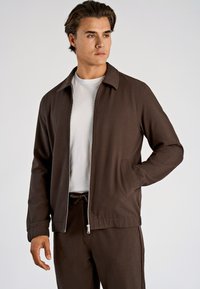 Bruine jas met rits en kraag, met een gladde textuur en twee zijzakken. Draagt over een wit T-shirt, gecombineerd met bijpassende broek.