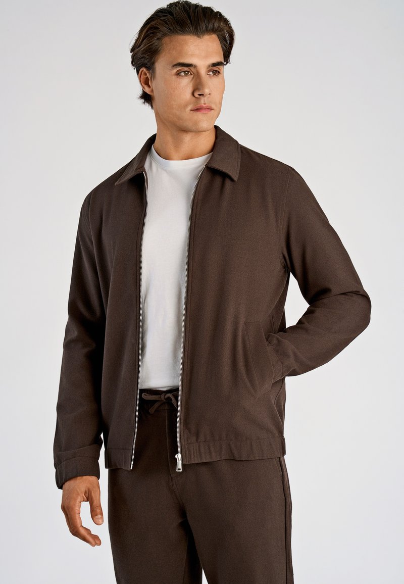 Bruine jas met rits en kraag, met een gladde textuur en twee zijzakken. Draagt over een wit T-shirt, gecombineerd met bijpassende broek.