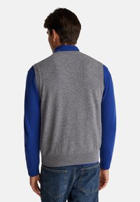 Szary sweter w kamizelce z dzianiny o gładkiej fakturze, noszony na niebieskiej koszuli z długim rękawem. Posiada prążkowany dół i bezrękawnikowy krój.