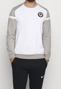 Felpa a maniche lunghe bianca e grigia con un logo rotondo sul lato sinistro del petto, caratterizzata da un colletto e polsini a costine. Abbinata a pantaloni neri.