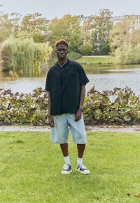 Chemise noire à manches courtes avec tissu texturé, portée sur un t-shirt noir. Shorts en denim clair, chaussettes blanches et baskets noires. Fond de parc vert.