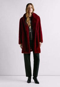 Cappotto rosso in pelliccia sintetica con ampio colletto, modello fino al ginocchio e due tasche laterali, indossato sopra pantaloni scuri con trama e stivali neri appuntiti.