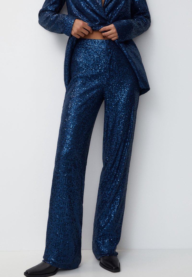 Personne portant un pantalon bleu foncé à sequins et un blazer assorti, ajustant l'ourlet du blazer, debout contre un mur clair.