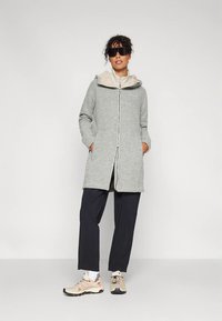 Kobieta o kręconych włosach, ubrana w czarne okulary przeciwsłoneczne, szary płaszcz na zamek, czarne spodnie, białe skarpetki oraz beżowe sneakersy, stojąca ze złożonymi w kieszeniach rękami.