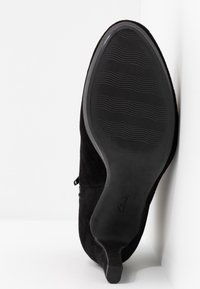 Semelle d'une botte en daim noir à petit talon carré, montrant une semelle extérieure en caoutchouc texturée avec un motif ondulé.