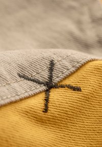 Tissu en coton avec des sections grises et jaunes, présentant des coutures texturées visibles et un détail de croix noire brodé à la couture.