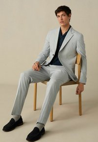 Helehall tekstureeritud ülikond, millel on ühe nupuga jakk ja sobivad püksid, kombineeritud tumesinise mustrilise särgi ja mustade loafers-kingadega.