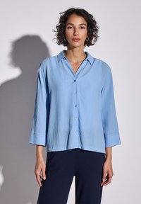 Camicia azzurra chiara con tessuto textured, maniche lunghe, colletto a punta e vestibilità leggermente ampia. Abbinata a pantaloni scuri.
