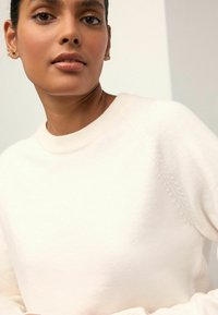 Sweatshirt crème clair avec un col rond et des manches raglan, présentant une texture douce et un détail de couture subtil le long des épaules.