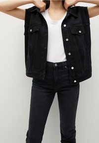Chaleco de mezclilla negro sin mangas con bolsillos frontales, detalles de botones plateados, usado sobre una camiseta blanca y combinado con jeans negros de cintura alta.