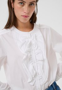 Jonge vrouw in een witte blouse met ruches aan de voorkant en een blauwe jeans, poserend tegen een effen lichte achtergrond.