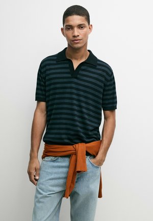 Ung mand iført en sort og mørkeblå stribet poloshirt med en rustfarvet sweater bundet om livet og lyseblå jeans.