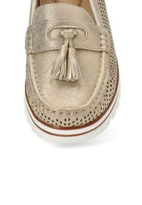 Mocasín metálico dorado con diseño perforado; presenta un acento de borlas y suela blanca con ribete marrón. Textura suave.
