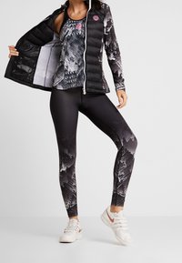 Giacca a vento nera sopra un top a fantasia e leggings neri lucidi, con un design montano. Sneakers bianche completano il look.