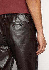 Pantalones de piel marrón con una textura suave, diseño ajustado y un bolsillo trasero con detalle de botón negro.