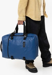 Blå duffelväska gjord av hållbart material, med svarta detaljer, en dragkedja upptill och justerbara remmar. Dimensionerna tyder på ett rymligt inre.