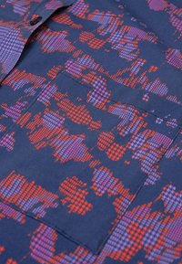 Marineblauwe stof met een gestructureerd patroon van paarse en rode geometrische vormen. Bevat een zichtbare zak en knoopdetail. Zachte, geweven stof.