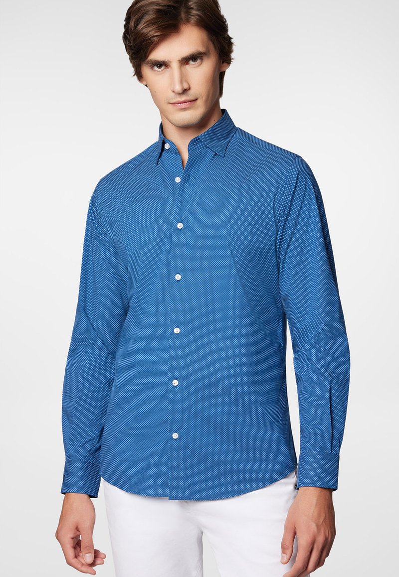 Niebieska koszula z długim rękawem i wzorem geometrycznym, z szerokim kołnierzem. Gładka tkanina z białymi guzikami. Noszona z białymi spodniami.