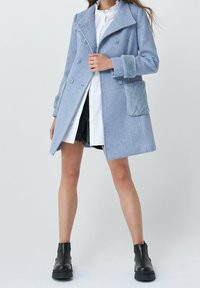 Donna che indossa un cappotto doppiopetto azzurro chiaro con polsini e tasche in pelliccia sintetica, stivaletti neri alla caviglia e un vestito camicia bianco.