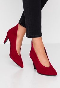 Zapatos de tacón alto de ante rojo con puntera puntiaguda y laterales recortados, con un delgado tacón de aguja y suela negra.