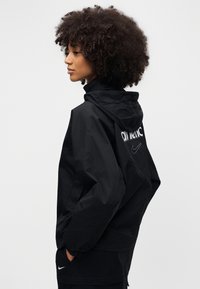 Veste coupe-vent noire avec capuche à cordon de serrage, poignets élastiques et logo au dos. Le tissu a une texture mate.