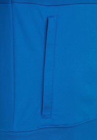 Modrý sportovní materiál s hladkou texturou, s vertikální kapsou na boku a žebrovaným elastickým pasem dole.