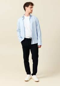 Camisa de botones azul claro sobre una camiseta blanca, combinada con pantalones de chándal oscuros y zapatillas blancas, de pie sobre un fondo claro.