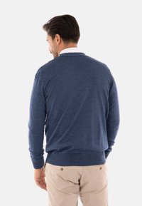 Maglione blu navy in tessuto lavorato a maglia, collo a giro, polsini e orli a costine, indossato sopra pantaloni chiari, visto da dietro.
