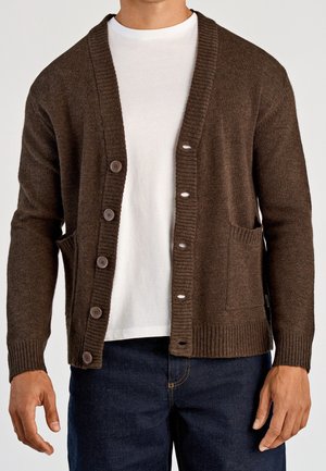 Mężczyzna w brązowym kardiganie na guziki, nałożonym na białą koszulkę i ciemnoniebieskie dżinsy, stojący z rękami swobodnie opuszczonymi wzdłuż ciała.