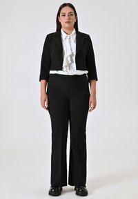 Zwarte cropped blazer over een witte blouse met rimpel-kraag, gecombineerd met zwarte flared broek en stevige zwarte laarzen. Eenvoudige, professionele look.