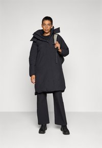 Zwarte geïsoleerde parka met een capuchon, voorzijde met ritssluiting en zijzakken. Het materiaal lijkt dik en gestructureerd, gecombineerd met zwarte broeken en schoenen.
