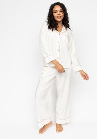 Weißes Schlafanzugset aus glattem Stoff; umfasst langärmeliges, knöpfbares Hemd mit Brusttasche und weit geschnittene Hose mit braunem Piping.