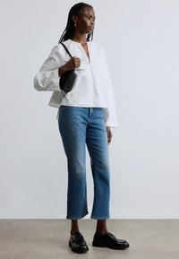 Fehér, laza szabású blúz V-nyakkal, kék farmer rövidnadrág és fekete bőr cipő. Texturált fekete kézitáskát tart a kezében.