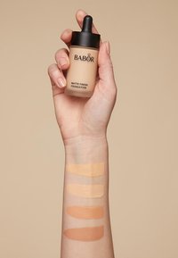 Babor foundation med matt finish i en pipettflaska som hålls i en hand. Fyra foundationprover i olika beige nyanser på en underarm.