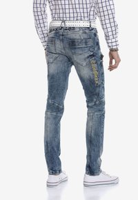 Helle blaue Denim-Jeans mit einem verwachsenen Design, mehreren Taschen und gestickten Akzenten. Kombiniert mit einem weißen Gürtel und weißen Sneakern.