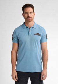 Ljusblå polo skjorta i bomull, med klassisk krage, knappstängning, broderade lappar på bröstet och ärmen, samt korta ärmar.