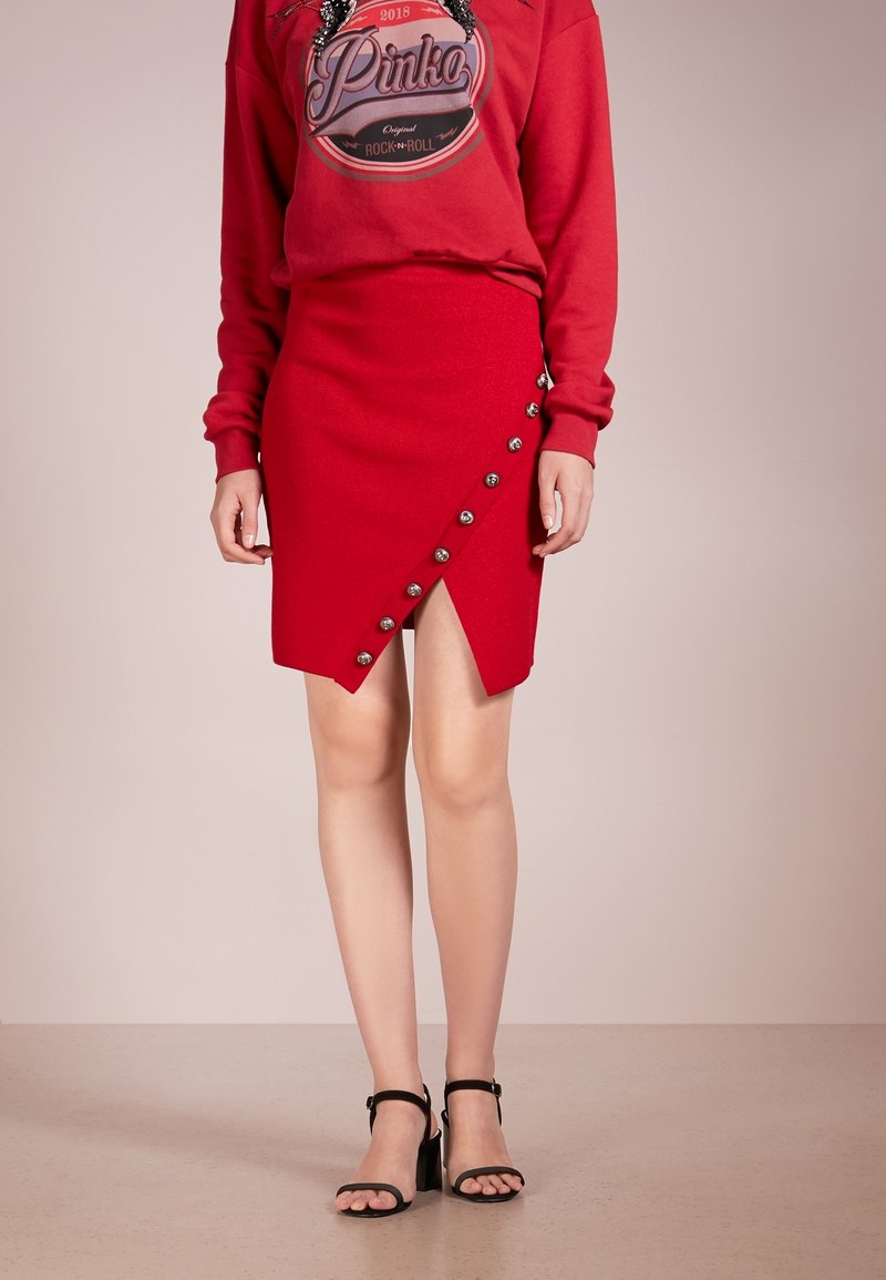 Jupe rouge avec un ourlet asymétrique, ornée de boutons métalliques le long de la couture diagonale. Texture de tissu lisse et design ajusté.