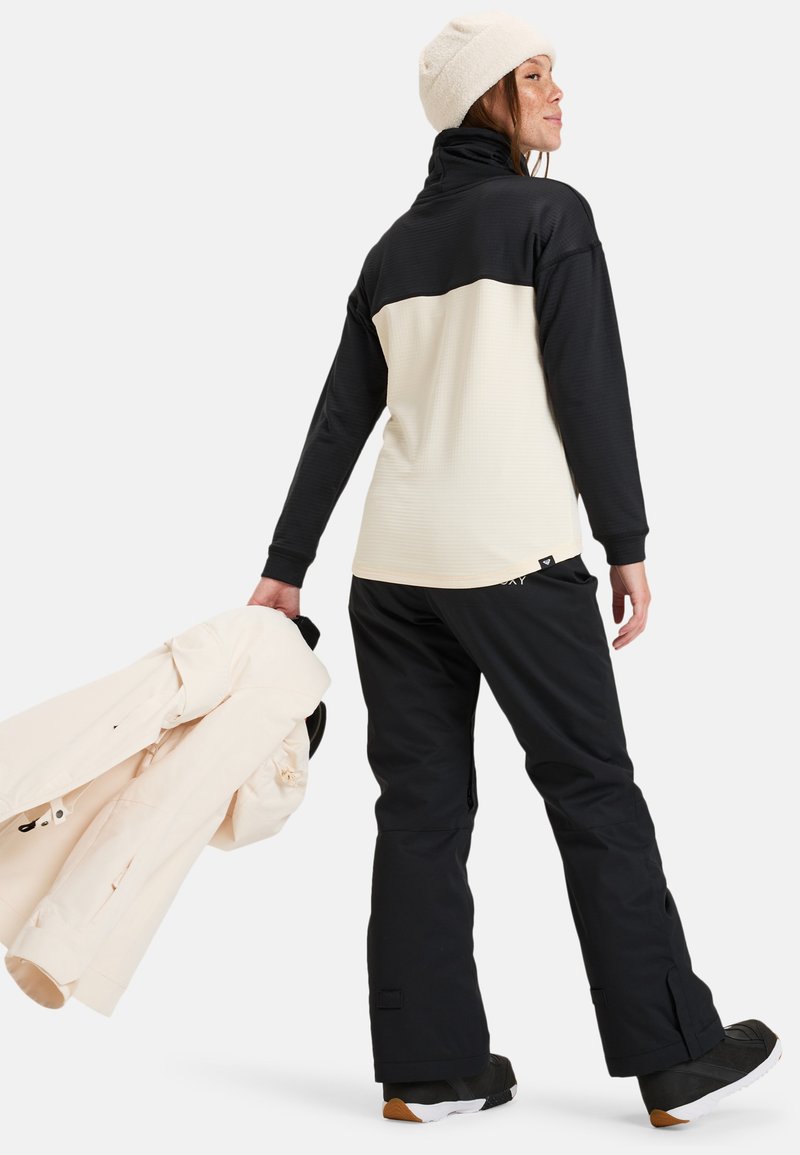 Zwarte en crème thermoshirt met hoge kraag en textuurdesign; gecombineerd met zwarte broek en een lichtcrème jack vasthoudend. Muts op het hoofd.
