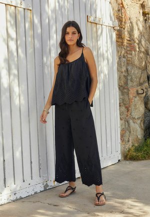 Top nero senza maniche con orlo ondulato e dettagli ricamati, abbinato a pantaloni neri a gamba larga con motivi decorativi e tasche.