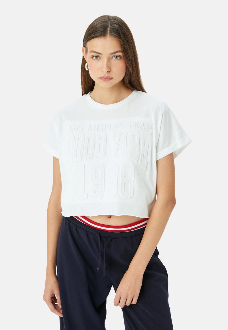 Witte, cropped T-shirt met reliëftekst "LOS ANGELES TEAM GOOD VIBE 1998." Korte mouwen en ronde halslijn; van zacht katoen.