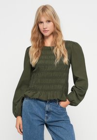 Blouse smockée vert foncé avec des manches bouffantes et un ourlet froncé, associée à un jean en denim bleu clair. Texture de tissu lisse, design décontracté.
