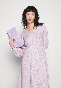 Vestido floral em lavanda com mangas bufantes longas e decote em V. Acompanha uma carteira suave em lavanda combinando. O tecido é leve com um estampado delicado.